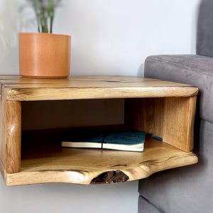 May include: A wooden floating shelf with a natural wood finish. The shelf has a single open shelf and a curved front edge. A small potted plant and a book are on the shelf.