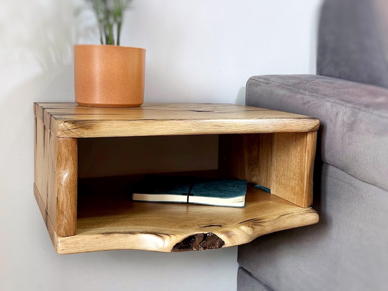 May include: A wooden floating shelf with a natural wood finish. The shelf has a single open shelf and a curved front edge. A small potted plant and a book are on the shelf.