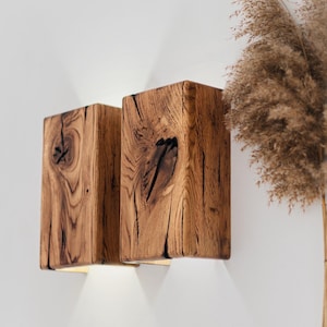 Pode incluir: Duas arandelas de parede de madeira com acabamento natural de grão de madeira. As arandelas são retangulares e emitem uma luz quente de baixo. A madeira tem nós e padrões escuros. Iluminação decorativa para casa.