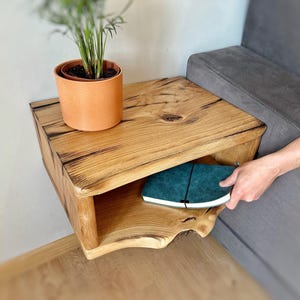 Könnte beinhalten: Ein rustikaler Nachttisch aus Holz mit natürlicher Holzmaserung. Der Tisch hat ein eingebautes Regal und eine kleine Topfpflanze oben drauf. Ein tealfarbenes Notizbuch wird auf das Regal gelegt.