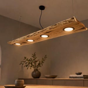 Pode incluir: Uma luminária pendente linear com uma tábua de madeira natural de borda irregular suspensa por cabos finos. A viga de madeira está equipada com quatro focos LED circulares embutidos que emitem um brilho quente voltado para baixo.