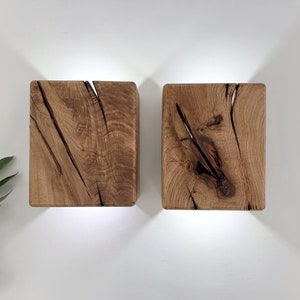 May include: Two square wooden wall sconces with a natural wood finish. The sconces have a rustic, reclaimed look with knots and grain patterns. The sconces are lit from the inside, casting a warm glow.