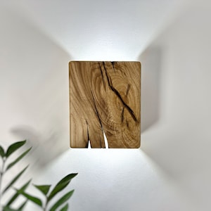 May include: A square wooden wall sconce with a natural wood finish and a light source shining through the cracks in the wood.