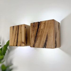 May include: Two rustic wooden wall sconces with a natural wood finish and a light brown color. The sconces are square shaped and have a rough, textured surface.