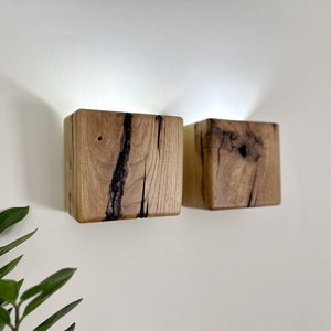 May include: Two square wooden wall sconces with a light source inside. The sconces are made of natural wood with dark brown streaks and knots.