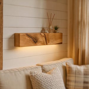Puede incluir: Una estantería flotante de madera con acabado natural está montada en una pared blanca. La estantería tiene luz integrada y está decorada con un difusor de vidrio, cañas y una pequeña planta en maceta. La estantería está encima de un sofá beige.