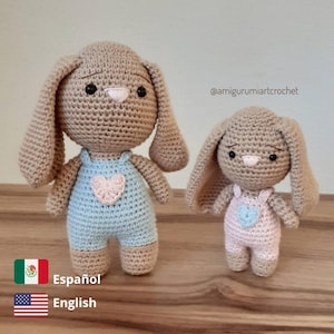 Puede incluir: Dos peluches de conejo de crochet, uno más grande que el otro. El conejo más grande lleva un mono azul con un corazón rosa, y el conejo más pequeño lleva un mono rosa con un corazón rosa. Ambos conejos tienen pelo marrón y ojos negros.