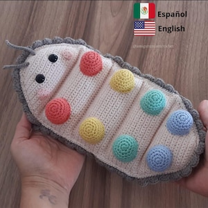 Puede incluir: Un juguete de oruga de crochet con un cuerpo gris y círculos de colores. La oruga tiene una cara con dos ojos negros y una nariz rosa.