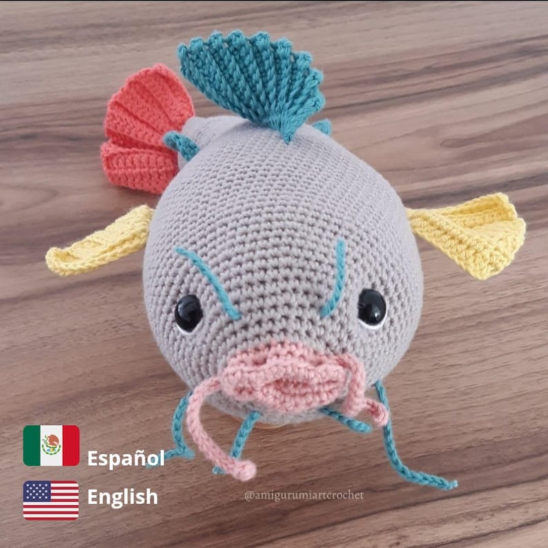 May include: A crocheted grey catfish toy with yellow, orange and blue fins. The catfish has black eyes and a pink mouth.