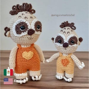 Puede incluir: Dos peluches de perezoso tejidos a crochet, uno más grande que el otro, ambos con un mono naranja con un diseño de corazón. El perezoso más grande tiene el pelo marrón y el perezoso más pequeño tiene el pelo marrón en la parte superior de la cabeza. Los peluches están de pie sobre una superficie de madera. @amigurumiartcrochet