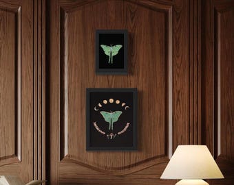May include: Two framed art prints featuring a green moth design on a black background. The top print shows a single moth, while the bottom print includes crescent moons, a mushroom, and a leafy border. The frames are black, and the artwork hangs on a wood-paneled wall.