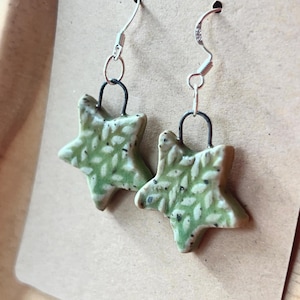 May include: A pair of star-shaped earrings. Each earring features a light green, textured star with a leaf pattern and black speckles. The earrings hang from silver-colored ear wires.