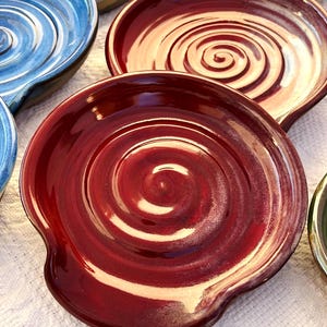 May include: A collection of handmade ceramic spoon rests in various colors. The spoon rests feature a spiral design in the center and a curved edge for holding utensils. Colors include deep red, blue, and tan.