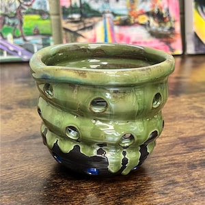 May include: A handmade ceramic vessel with a layered design. The top and middle sections are glazed in a glossy green, with evenly spaced holes. The bottom section is dark brown with blue accents.