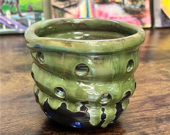 May include: A handmade ceramic vessel with a layered design. The top and middle sections are glazed in a glossy green, with evenly spaced holes. The bottom section is dark brown with blue accents.