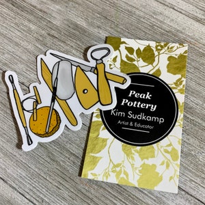 May include: A sticker with a drawing of pottery tools, including a wooden handle, a metal scraper, a wooden tool, and a sponge. The sticker is on a wooden surface next to a business card that reads "Peak Pottery Kim Sudkamp Artist & Educator".