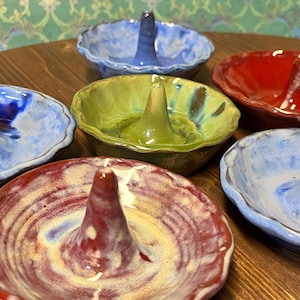 May include: A collection of handmade ceramic ring holders in various colors, including blue, green, and red. Each holder features a central cone for ring placement and a shallow dish base. The ring holders are displayed on a wooden surface.