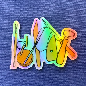 May include: A rainbow holographic sticker featuring a collection of pottery tools, including a wooden paddle, a metal scraper, a small wooden tool, a metal needle tool, and a wooden handle with a metal loop. The tools are arranged around a small brown clay disc.