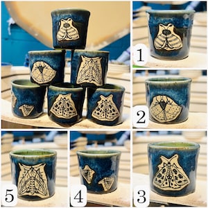 Può includere: Una collezione di bicchierini in ceramica fatti a mano. Ogni bicchiere blu scuro presenta un esclusivo disegno di falena in crema e nero. I bicchieri hanno una finitura lucida e un bordo verde. I bicchierini sono impilati.