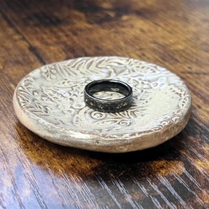 May include: A black ring with a textured band and gold-colored accents rests on a small, round, off-white ceramic dish with a floral pattern. The dish is on a wooden surface.