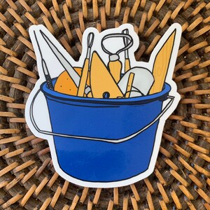 May include: A blue bucket filled with various sculpting tools, including a wooden paddle, a metal scraper, and a few paintbrushes. The bucket is sitting on a woven wicker surface.