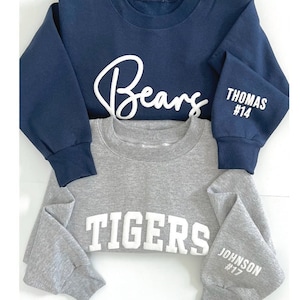 Puede incluir: Dos sudaderas, una azul marino con "Bears" y "THOMAS #14" en las mangas, y otra gris con "TIGERS" y "JOHNSON #17" en las mangas.