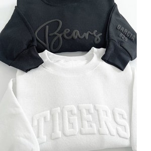 Peut inclure: Deux sweatshirts, un noir et un blanc, sont empil&eacute;s. Le sweat-shirt noir porte l'inscription "Bears" en relief et "GARCIA #29" sur la manche. Le sweat-shirt blanc porte l'inscription "TIGERS" en relief. Col rond.