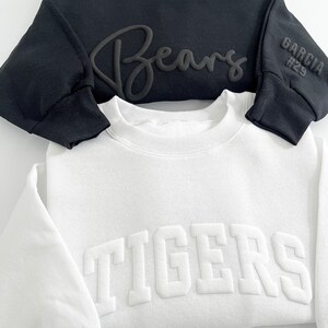 Puede incluir: Dos sudaderas, una negra y una blanca. La sudadera negra tiene "Bears" escrito en cursiva en la parte delantera y "Garcia #29" en la manga. La sudadera blanca tiene "TIGERS" escrito en letras grandes y en negrita en la parte delantera.