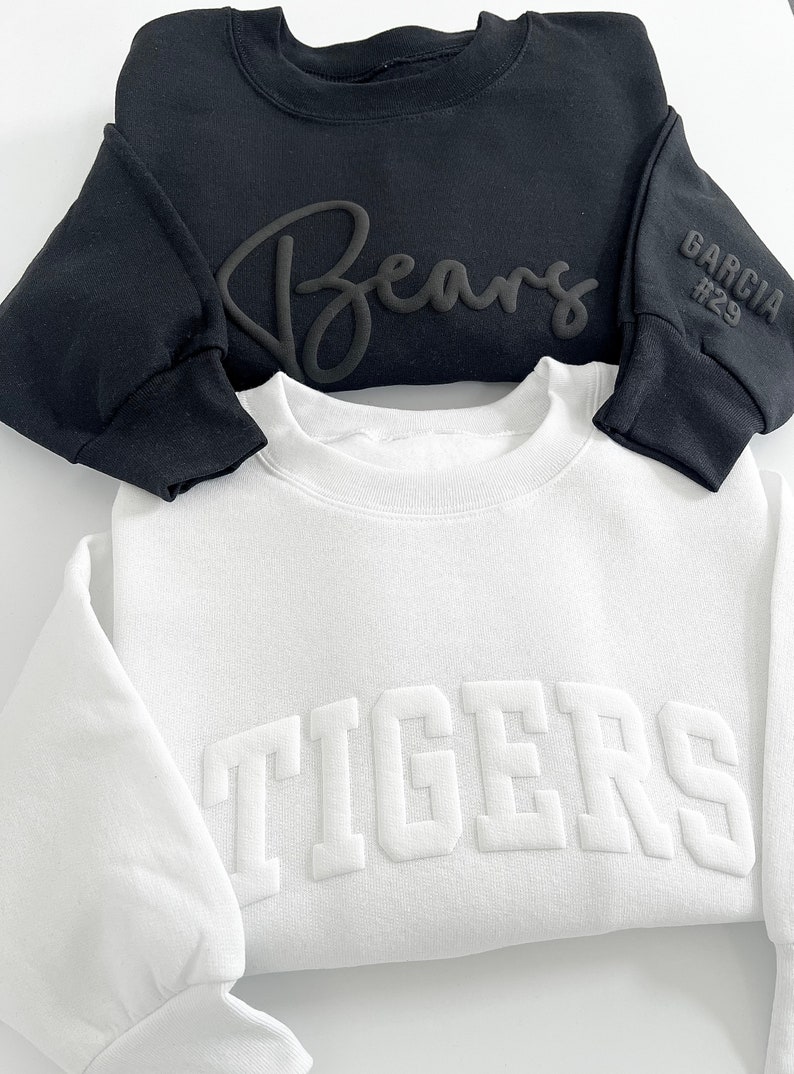 Puede incluir: Dos sudaderas, una negra y una blanca. La sudadera negra tiene "Bears" escrito en cursiva en la parte delantera y "Garcia #29" en la manga. La sudadera blanca tiene "TIGERS" escrito en letras grandes y en negrita en la parte delantera.