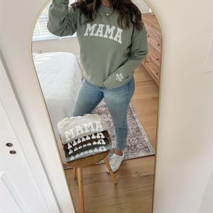 May include: A stack of four sweatshirts in different colors, each with the word "MAMA" printed in white on the front. The sweatshirts are folded and stacked on a wooden stool. The sweatshirts are in the colors beige, brown, gray, and green.