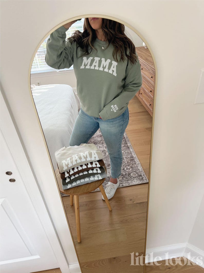 May include: A stack of four sweatshirts in different colors, each with the word "MAMA" printed in white on the front. The sweatshirts are folded and stacked on a wooden stool. The sweatshirts are in the colors beige, brown, gray, and green.