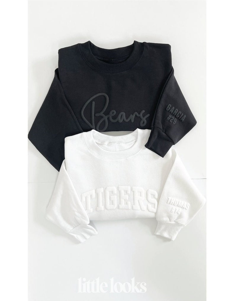 Peut inclure: Deux sweat-shirts col rond, un noir et un blanc, sont pr&eacute;sent&eacute;s. Le sweat-shirt noir porte l'inscription "Bears" en &eacute;criture cursive et "GARCIA #29" sur la manche. Le sweat-shirt blanc affiche "TIGERS" et "THOMAS #14" sur la manche. La marque "little looks" est en bas.