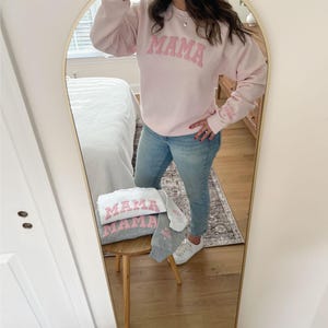 May include: A pink sweatshirt with the word "MAMA" in pink letters on the front. The sweatshirt is being worn by a person in a mirror reflection. There is a gray sweatshirt with the word "MAMA" in pink letters on the front on a stool in the reflection.