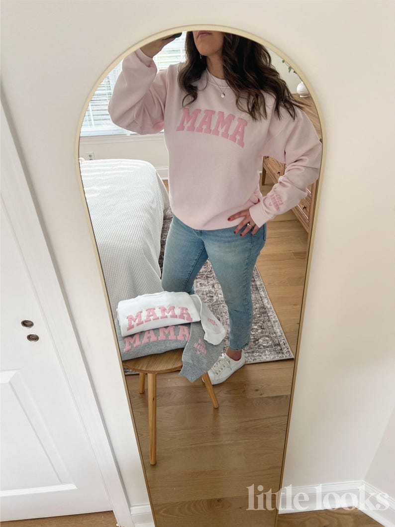 May include: A pink sweatshirt with the word "MAMA" in pink letters on the front. The sweatshirt is being worn by a person in a mirror reflection. There is a gray sweatshirt with the word "MAMA" in pink letters on the front on a stool in the reflection.