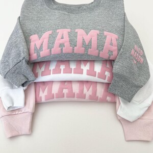 May include: A stack of three sweatshirts with the word "MAMA" printed in pink on each. The top sweatshirt is gray, the middle sweatshirt is white, and the bottom sweatshirt is pink. The sweatshirts are folded and stacked on top of each other. The text "little looks" is printed in white at the bottom of the image.