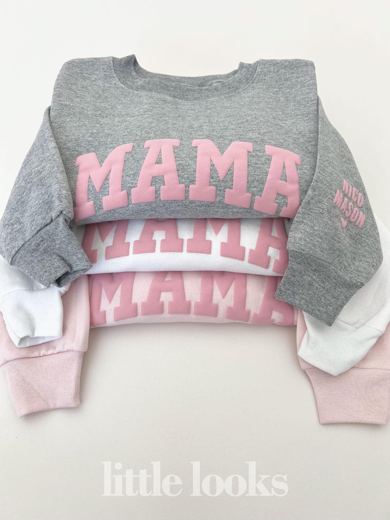 May include: A stack of three sweatshirts with the word "MAMA" printed in pink on each. The top sweatshirt is gray, the middle sweatshirt is white, and the bottom sweatshirt is pink. The sweatshirts are folded and stacked on top of each other. The text "little looks" is printed in white at the bottom of the image.
