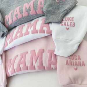 May include: Four pink sweatshirts with the word "MAMA" in pink raised letters. The sleeves of each sweatshirt have a different name and a heart printed on them. The names are "Nico Mason", "Luke Caleb", "Luca", and "Ariana". The text "little looks" is printed on the bottom of the image.
