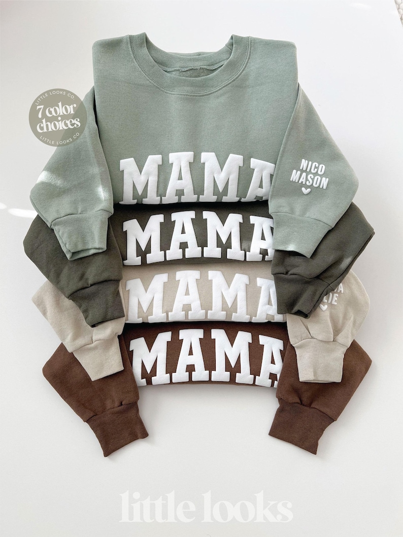 May include: A stack of green, brown, and beige sweatshirts with the word "MAMA" printed in white on the front. The sweatshirt on top has the words "NICO MASON" printed on the sleeve.