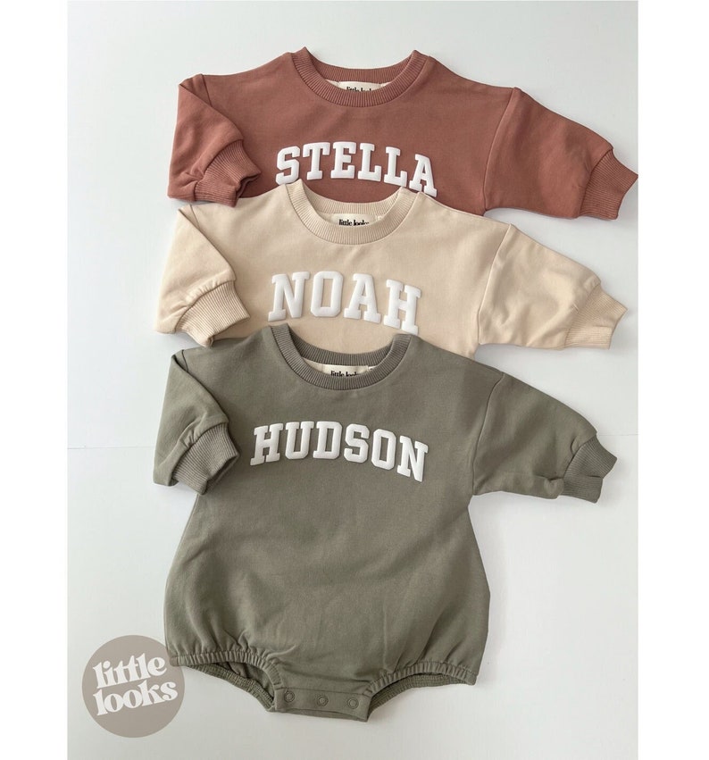 May include: Three baby onesies in different colors with white embroidered names: Stella, Noah, and Hudson. The onesies are long-sleeved and have a crew neck.