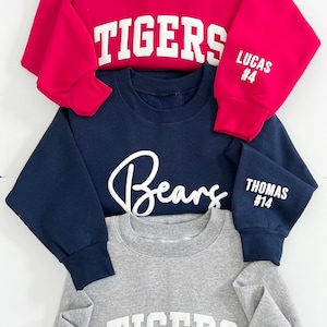 Peut inclure: Trois sweatshirts rouges, bleu marine et gris. Le rouge porte "TIGERS" devant et "LUCAS #4" sur la manche. Le bleu marine affiche "Bears" devant et "THOMAS #14" sur la manche. Le gris affiche "TIGERS" devant et "JOHNSON #17" sur la manche.