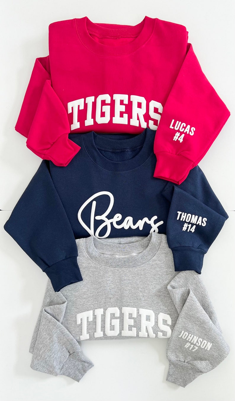 Peut inclure: Trois sweatshirts rouges, bleu marine et gris. Le rouge porte "TIGERS" devant et "LUCAS #4" sur la manche. Le bleu marine affiche "Bears" devant et "THOMAS #14" sur la manche. Le gris affiche "TIGERS" devant et "JOHNSON #17" sur la manche.