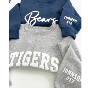 Puede incluir: Dos sudaderas, una azul marino con "Bears" escrito en cursiva blanca y "THOMAS #14" en la manga, y una gris con "TIGERS" en letras blancas y "JOHNSON #17" en la manga.