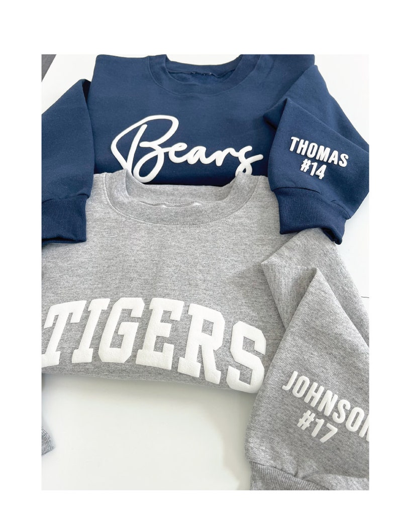 Puede incluir: Dos sudaderas, una azul marino con "Bears" escrito en cursiva blanca y "THOMAS #14" en la manga, y una gris con "TIGERS" en letras blancas y "JOHNSON #17" en la manga.