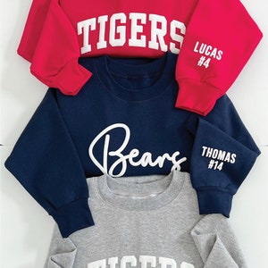 Puede incluir: Tres sudaderas con diferentes colores y dise&ntilde;os. La sudadera de arriba es roja con la palabra "TIGERS" en blanco en la parte delantera y "LUCAS #4" en la manga. La sudadera del medio es azul marino con la palabra "Bears" en blanco en la parte delantera y "THOMAS #14" en la manga. La sudadera de abajo es gris con la palabra "TIGERS" en blanco en la parte delantera y "JOHNSON #17" en la manga.