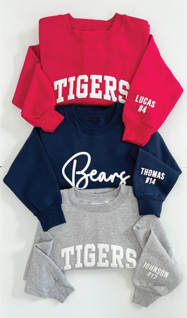 Puede incluir: Tres sudaderas con diferentes colores y dise&ntilde;os. La sudadera de arriba es roja con la palabra "TIGERS" en blanco en la parte delantera y "LUCAS #4" en la manga. La sudadera del medio es azul marino con la palabra "Bears" en blanco en la parte delantera y "THOMAS #14" en la manga. La sudadera de abajo es gris con la palabra "TIGERS" en blanco en la parte delantera y "JOHNSON #17" en la manga.