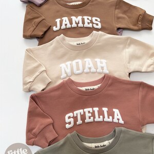 May include: Five personalized sweatshirts in different colors with names in white block letters. The names are Olivia, James, Noah, Stella, and Hudson. The sweatshirts are all from the brand "little looks".