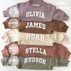 May include: Five crewneck sweatshirts in different colors with names embroidered in white on the front. The colors are dusty purple, coffee, cream, desert rose, and sage.