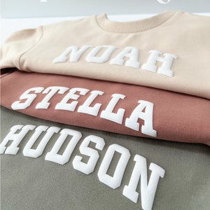 May include: Three sweatshirts stacked on top of each other. The top sweatshirt is beige with the name "NOAH" in white puffy letters. The middle sweatshirt is a dusty rose color with the name "STELLA" in white puffy letters. The bottom sweatshirt is a muted green color with the name "HUDSON" in white puffy letters.