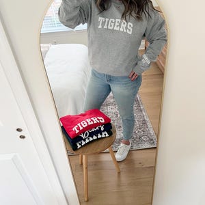 Puede incluir: Una pila de tres sudaderas con la palabra "TIGERS" impresa en blanco en cada una. Las sudaderas son roja, azul marino y gris. Las sudaderas est&aacute;n dobladas y apiladas en una mesa de madera.