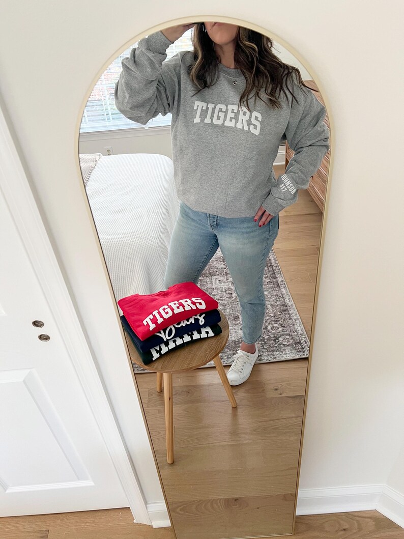 Puede incluir: Una pila de tres sudaderas con la palabra "TIGERS" impresa en blanco en cada una. Las sudaderas son roja, azul marino y gris. Las sudaderas est&aacute;n dobladas y apiladas en una mesa de madera.
